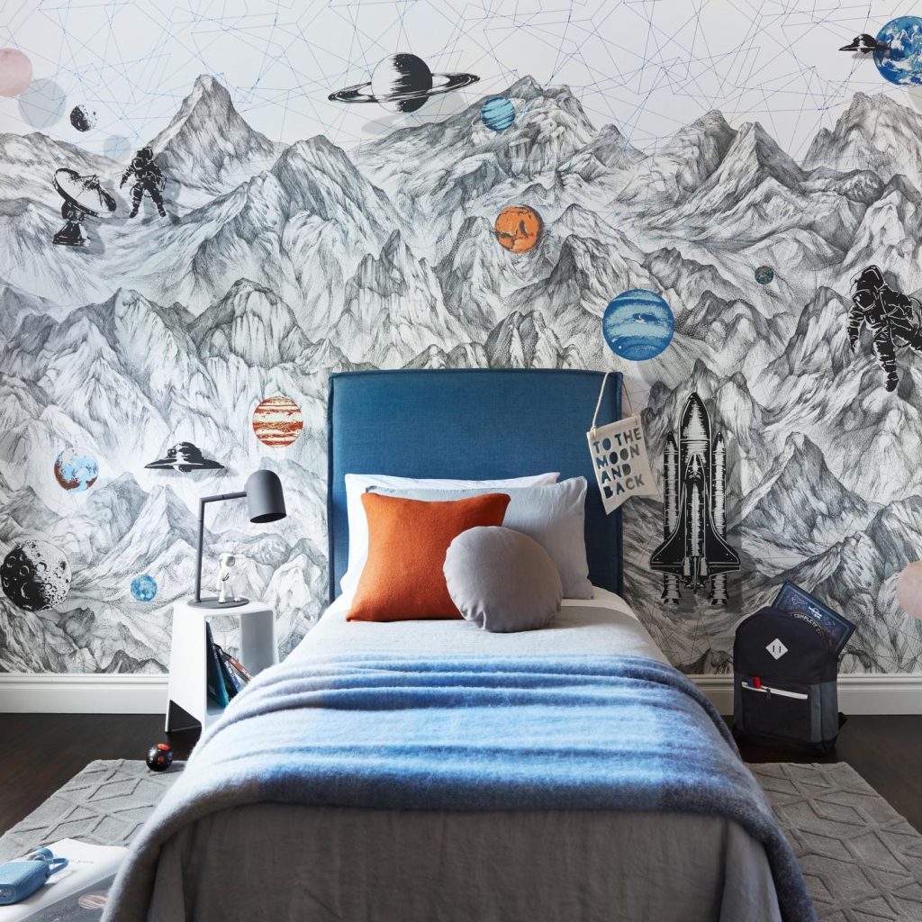 A child's bedroom with a space-themed mural on the wall, featuring mountains, planets, and rockets. A bed with blue and orange bedding is in the foreground. Bring Your Child's Dreams to Life with these decorating ideas for themed bedrooms.