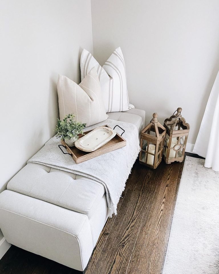 A cozy corner with a white bench adorned with elegant striped pillows, a tray, and flanked by two rustic wooden lanterns on a hardwood floor.