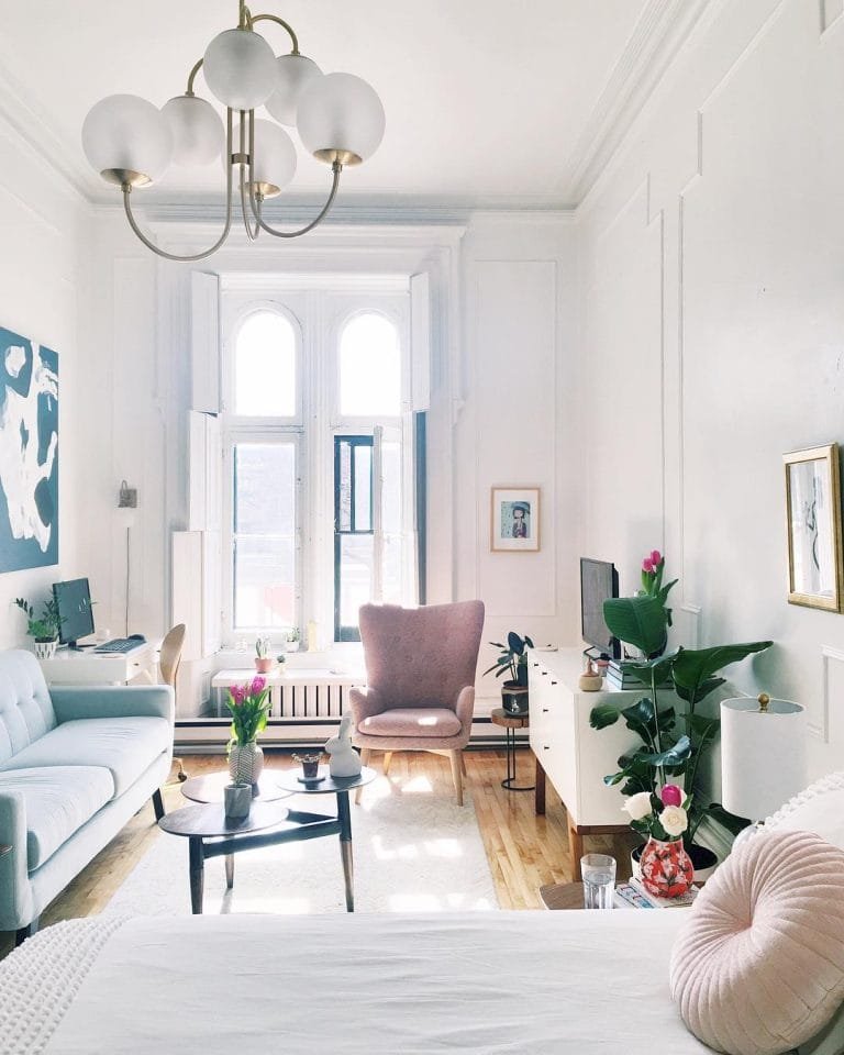 Bright, elegant living room in a studio apartment with large windows, white walls, various plants, and modern furniture including sofas and a chandelier.