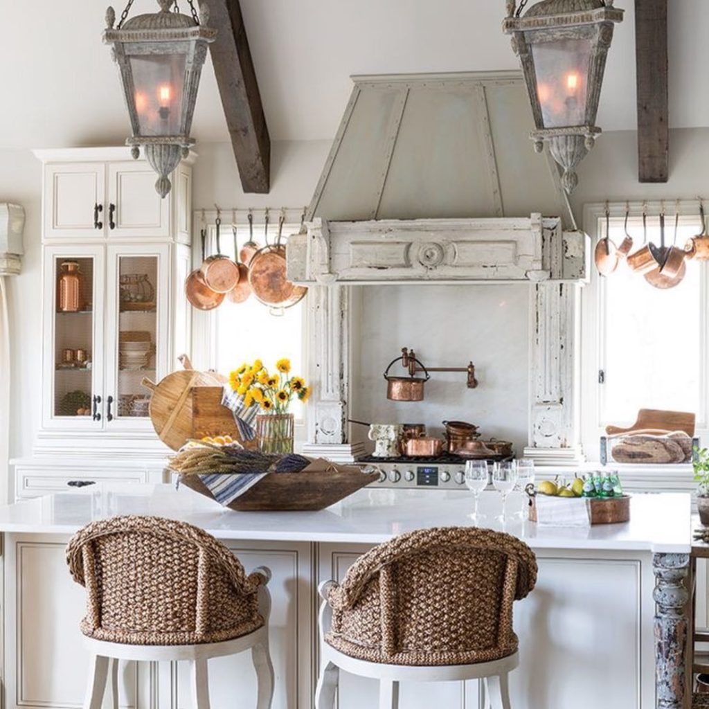 A bright farmhouse kitchen with a white island, wicker bar stools, hanging copper pots, two lantern lights, and a rustic range hood exemplifies the top 10 essential design elements for a French country style kitchen. Pineapples, lemons, and sunflowers add a vibrant touch to the counter.