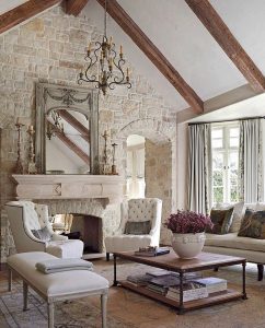 Elegant French country living room with stone walls, wooden beams, and a large mirror above the fireplace. Furnished with white chairs, a sofa, a coffee table with books, and a vase of flowers. Large windows with curtains balance functionality and beauty in this charming design.