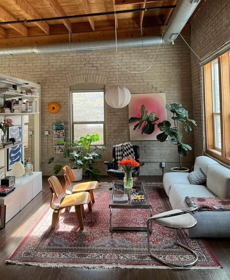 A cozy living room with a wooden ceiling, brick walls, a gray couch, two wooden chairs, several plants, a rug, bookshelves, and large windows letting in natural light—perfect for those wondering how to mix and match mid-century modern decor for a unique living room.