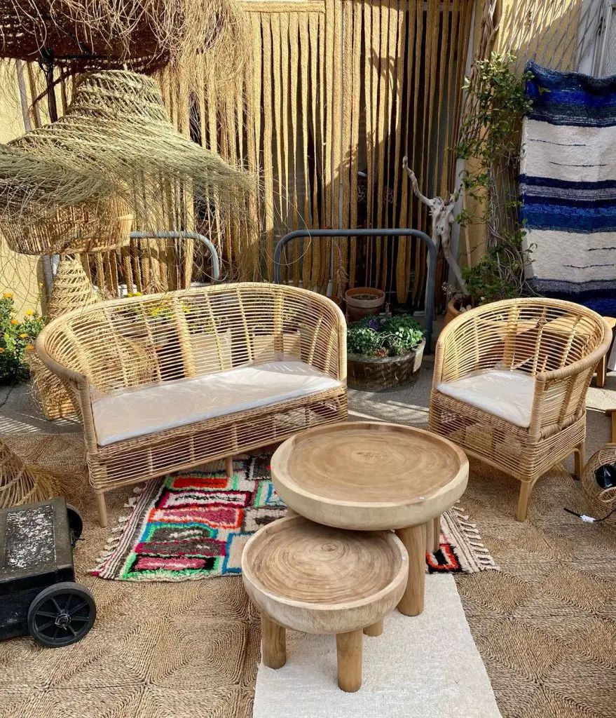A cozy Bohemian outdoor seating area with two rattan chairs, a sofa, and wooden tables on a patterned rug, surrounded by plants and rustic decor.