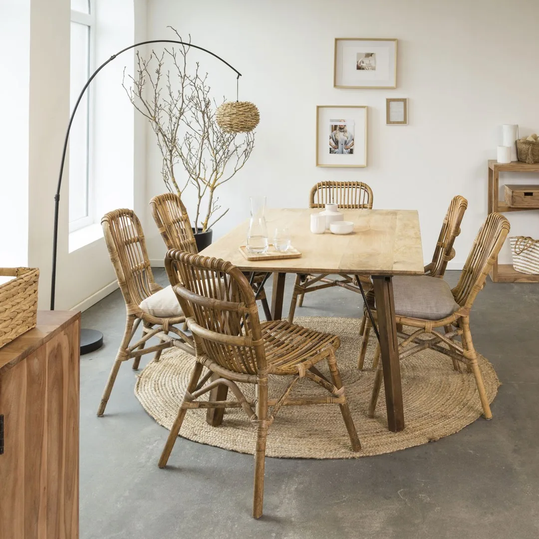 A rustic dining room with a wooden table, rattan chairs, and a circular rug, accented by a tall, arched floor lamp and simple wall art.