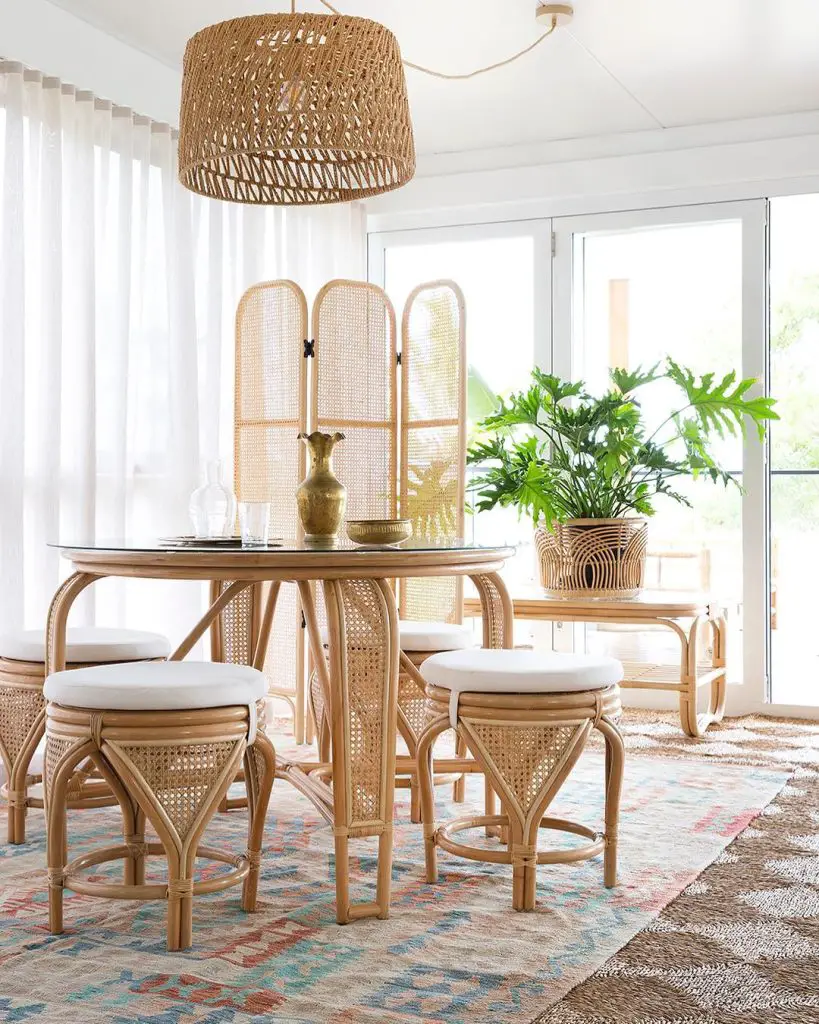 Bright, airy dining room with a round table, Bohemian rattan chairs, and a large woven light fixture, surrounded by large windows and a green plant.