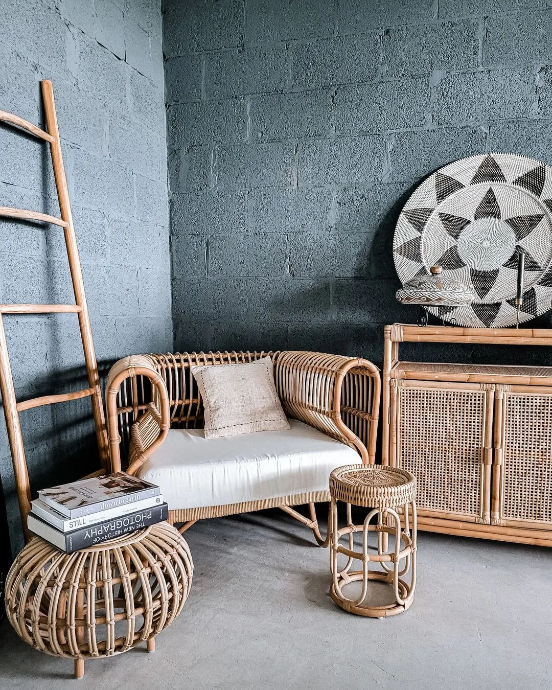 A cozy corner with Bohemian rattan bench, matching cabinet, and decorative items, set against a blue painted brick wall with a small ladder and art piece.