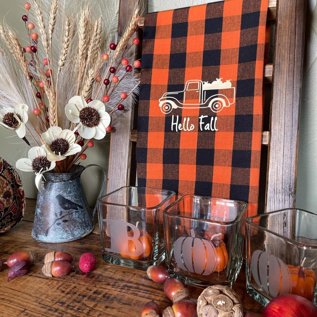 A fall-themed decoration features a sign reading "Hello Fall," a vase of fall flowers, and three glass jars with autumn designs. All are placed on a wooden surface surrounded by acorns and small pumpkins, showcasing creative ways to style with buffalo plaid for the perfect Fall Home Decor.