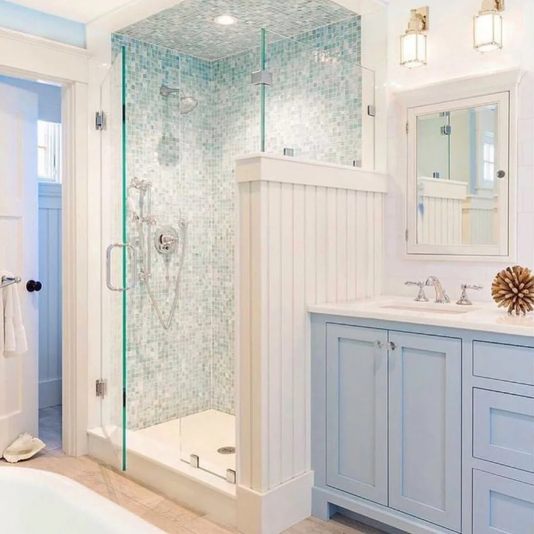 A modern coastal bathroom featuring a glass shower enclosure with turquoise tiles, a white bathtub, and light blue cabinetry.