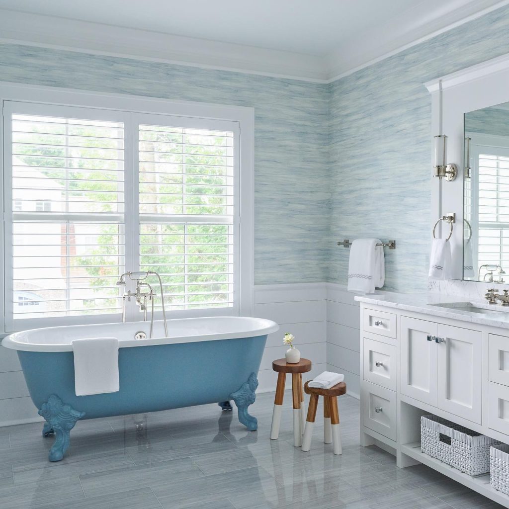 A spacious, modern bathroom with a blue freestanding bathtub, white cabinetry, and pale blue walls designed in coastal decor. Large windows provide natural light.