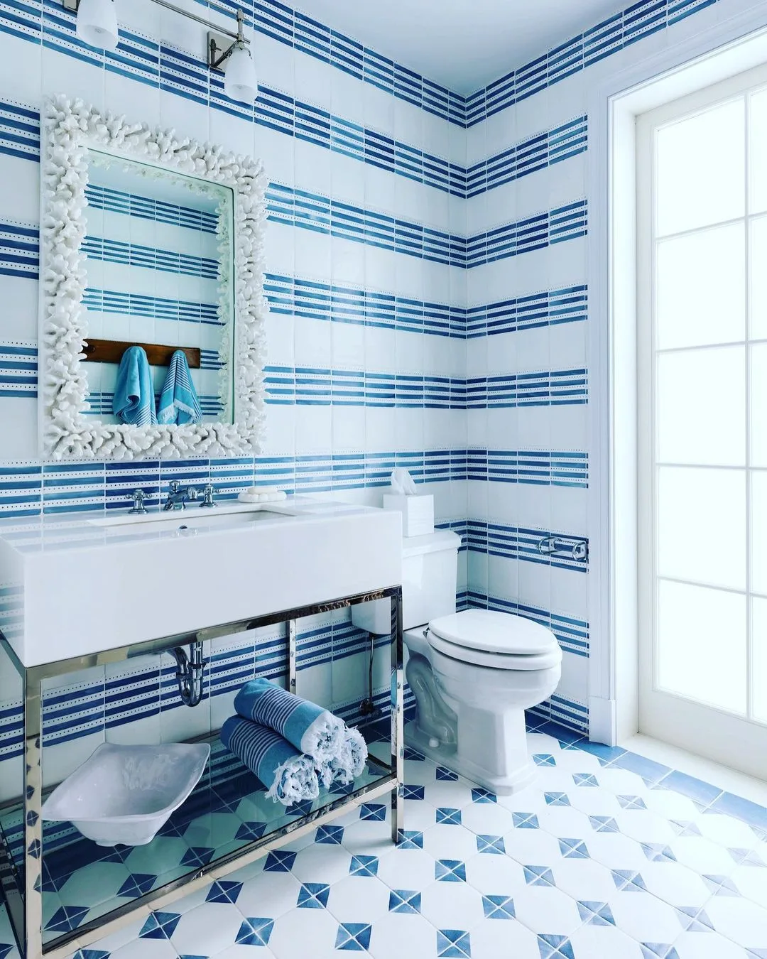 A coastal bathroom with blue striped wallpaper, geometric tiled floor, a white pedestal sink, mirror with a decorative frame, and a toilet next to a window.