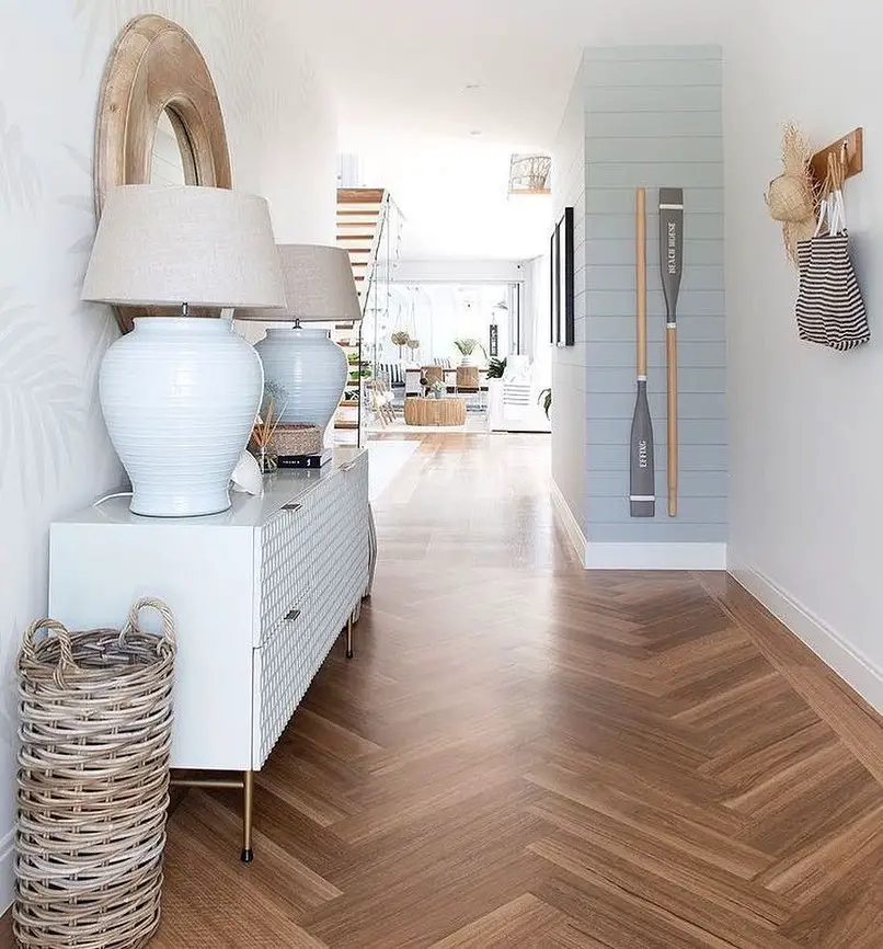 A hallway with wooden herringbone flooring features a white console table, two table lamps, a wicker basket, decorative oars on the wall, and a striped bag hanging on a hook. Stairs are visible in the background. Discover Coastal Entryway Decor: Tips and Tricks for Capturing the Perfect Coastal Vibe.