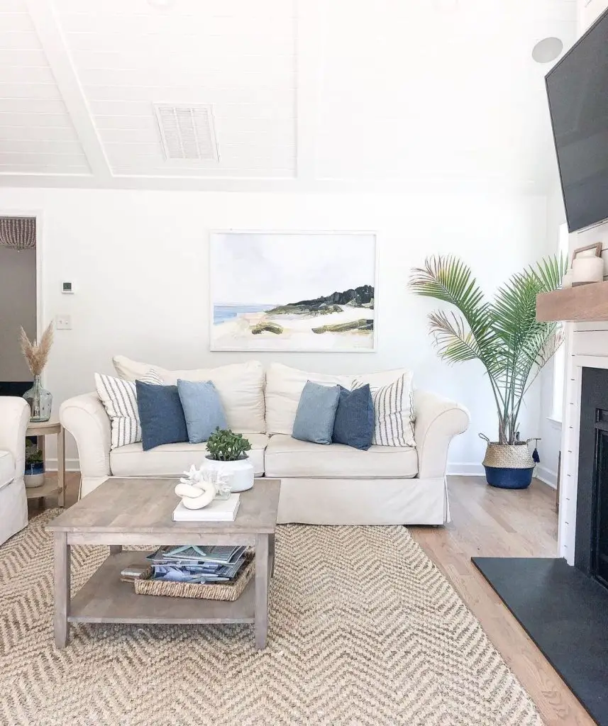 A bright living room with a white sofa adorned with blue cushions, a wooden coffee table, a textured rug, and a painting of a coastal scene on the wall.