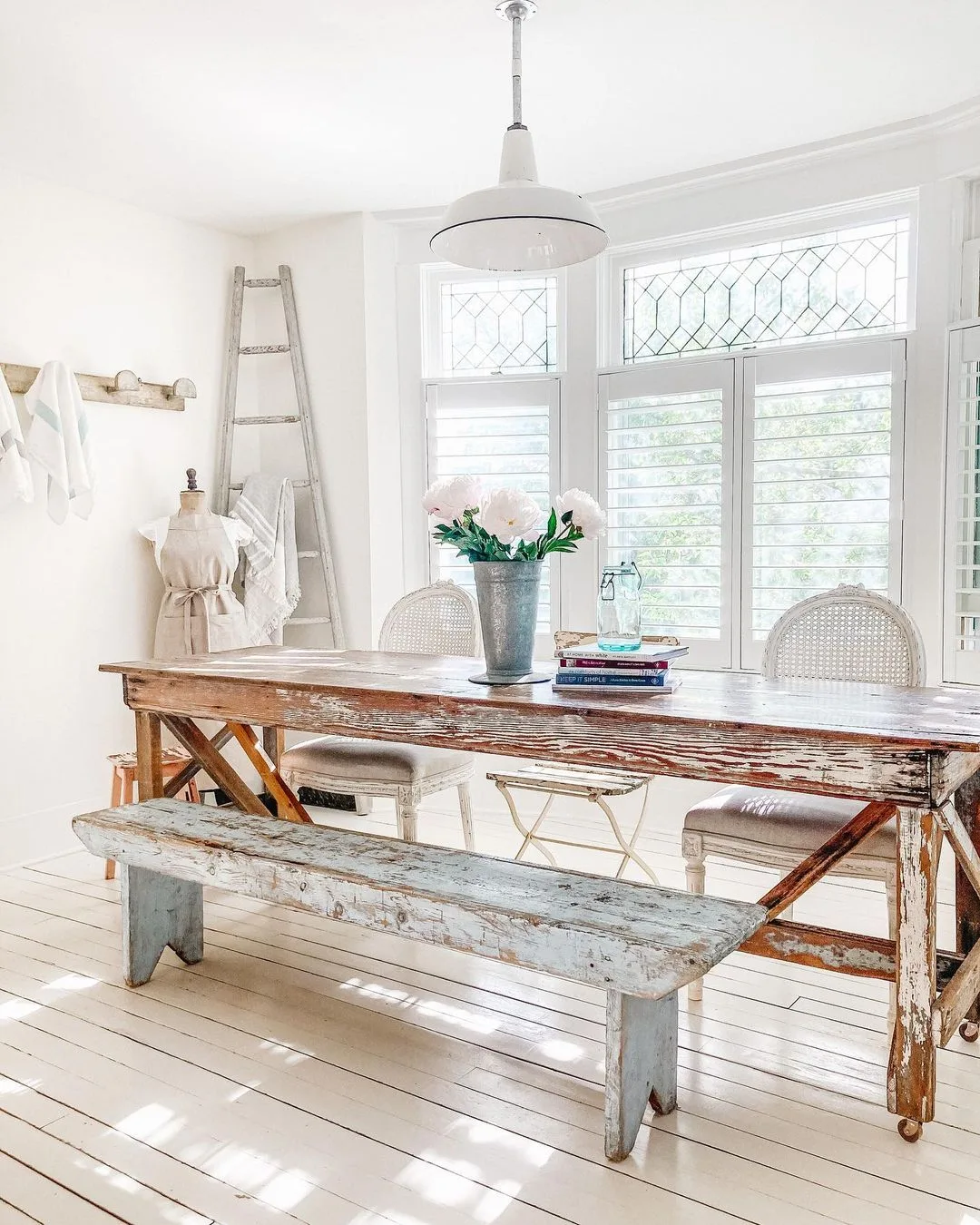 A rustic dining room showcases a wooden table, two wicker chairs, and a bench. A vase with flowers sits on the table. Against the white walls, you'll find a ladder, coat rack, and mannequin—a perfect blend of French Country living room design balancing functionality and beauty.