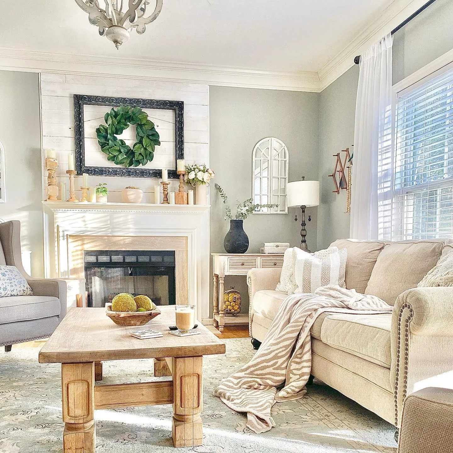 A bright French Country living room with a beige sofa, wooden coffee table, fireplace with a wreath above it, and a window with white curtains casting sunlight onto the room balances functionality and beauty seamlessly.