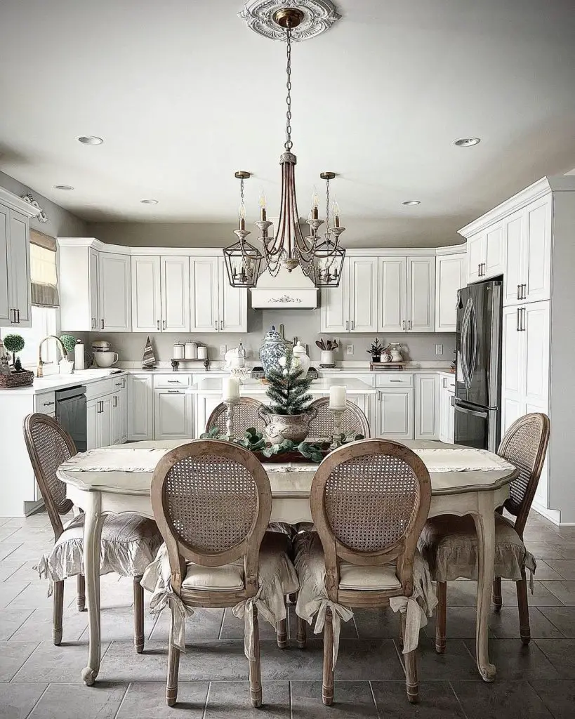 A bright, elegant kitchen features white cabinetry, a chandelier, and a dining table set for six with cushioned chairs. To create a warm and inviting French country dining room, a small decorative centerpiece adorns the table.