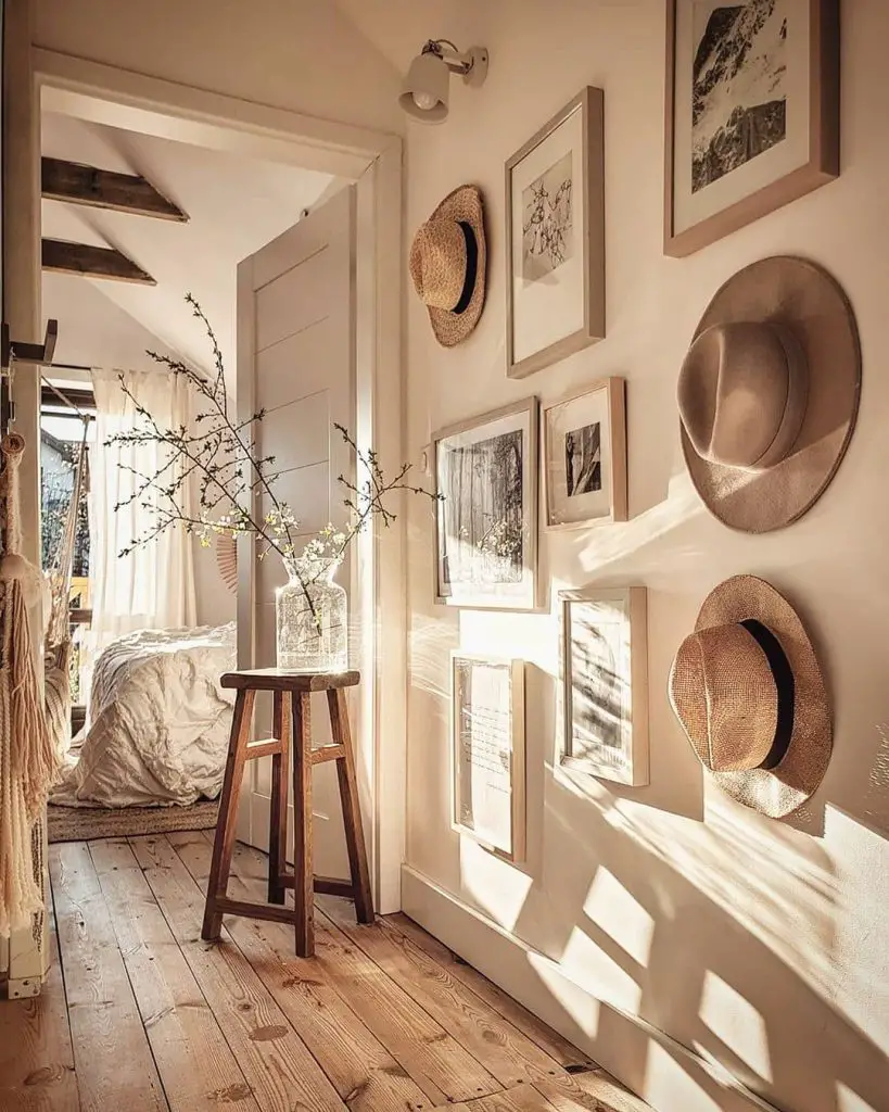 A sunlit hallway with a wooden floor, adorned with framed pictures and hats on the wall—a true testament to "The Art of the Hat: How to Curate a Hat Gallery Wall." A stool with a clear glass vase holding branches with flowers is placed near a partially open door.