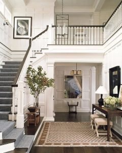 Elegant foyer with a dual staircase, white panel walls, and dark wood floors, featuring tasteful rustic-chic decor including indoor plants and a hanging pendant light.