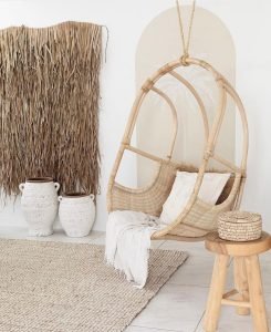 A cozy room featuring earthy and textured decor: a hanging wicker chair, a neutral-colored rug, and a wooden stool with a woven basket. The white decorative pots sit beside a natural woven divider against a white wall with a painted arch, perfect for styling your home with rattan decor.