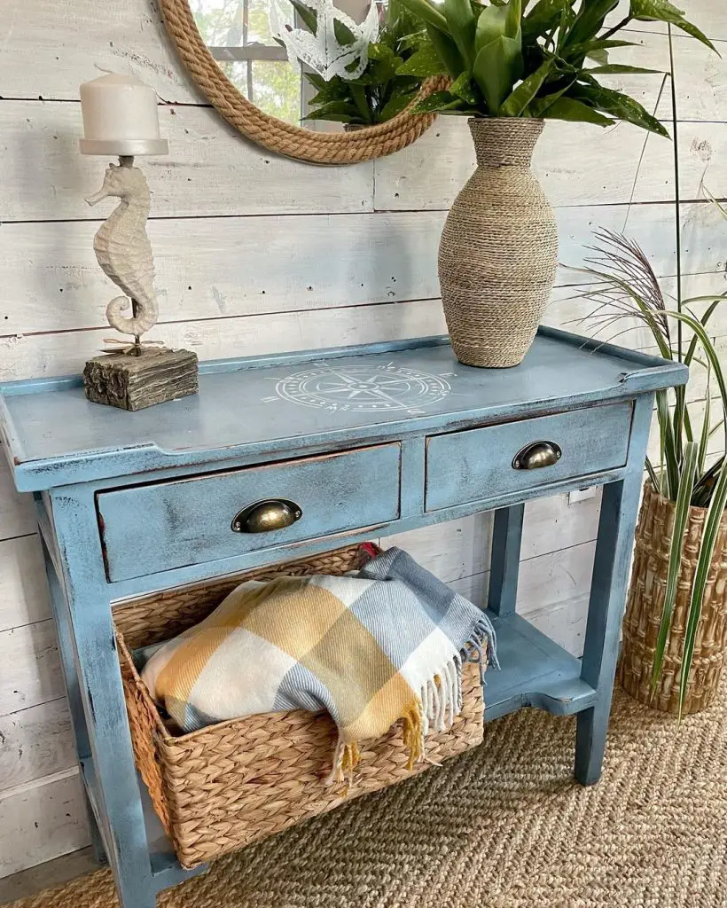 A rustic blue console table with two drawers, featuring a seashell design and bronze handles. On top, there's a seahorse-shaped candle holder and a woven vase with greenery. For those seeking tips on how to choose the perfect coastal console decor for your home, this setup offers ideal inspiration.
