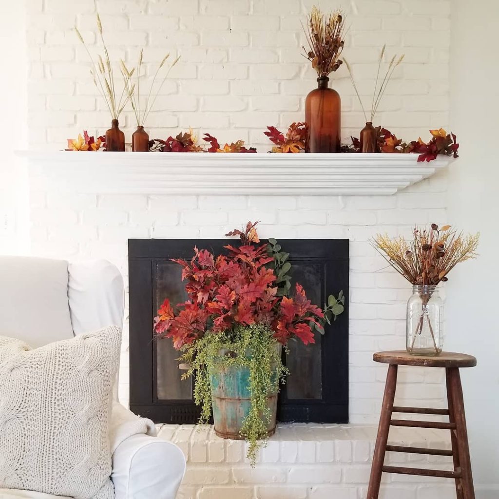 A white brick fireplace is decorated with fall-themed foliage, amber glass bottles, and dried flowers for a chic and sophisticated look. A potted plant sits in front of the fireplace, and a vase with twigs is placed on a stool, all forming neutral fall mantel decor ideas.