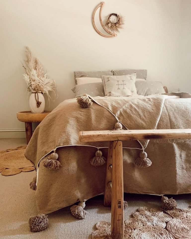 A cozy bedroom featuring a bed with a beige and textured coverlet adorned with tassels, a perfectly chosen rustic wooden bench at the foot of the bed, bedside vase with dried pampas grass, and crescent moon wall decor.