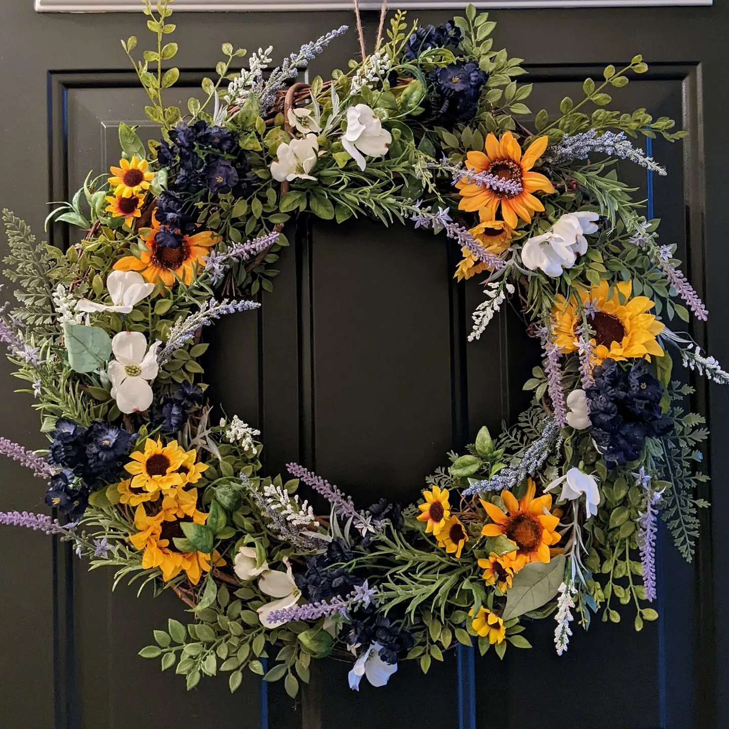A decorative wreath featuring a mix of yellow sunflowers, white magnolias, and purple flowers surrounded by green foliage is hanging on a dark-colored door. Learn how to make your own fall wreaths with natural materials and scents for a personalized touch.