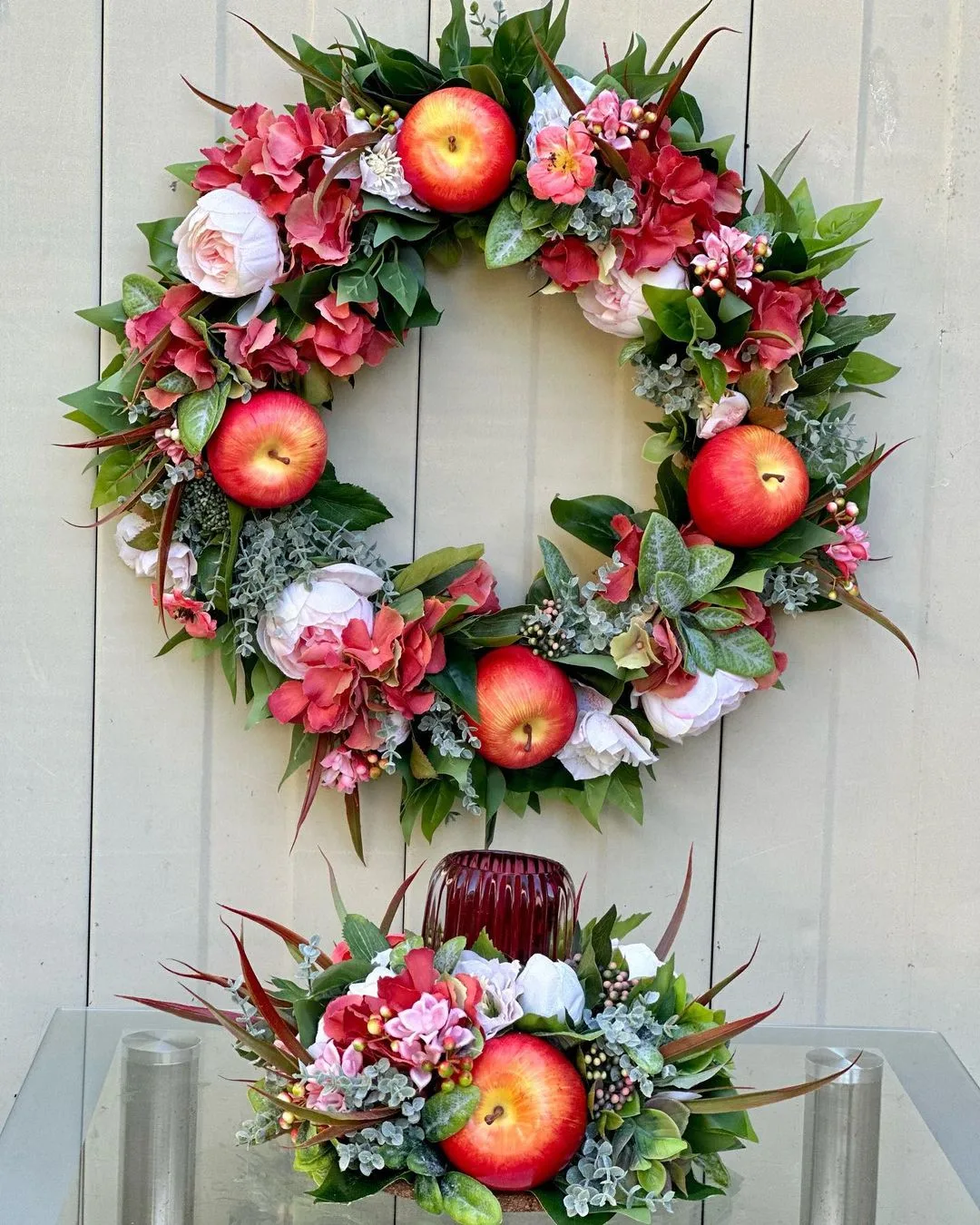A floral wreath with red apples, pink and white flowers, and greenery hangs on a wooden wall above a matching flower arrangement on a glass surface. Discover how to make your own fall wreaths with natural materials and scents to bring warmth to your home decor.
