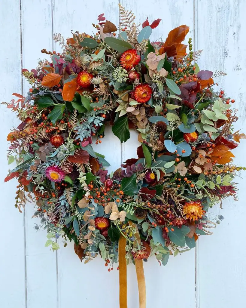 A wreath made of various autumn leaves, berries, and flowers, with additional green foliage and orange accents, is hung against a white wooden background. Learn how to make your own fall wreaths with natural materials and scents to bring this charming piece of seasonal decor to life.