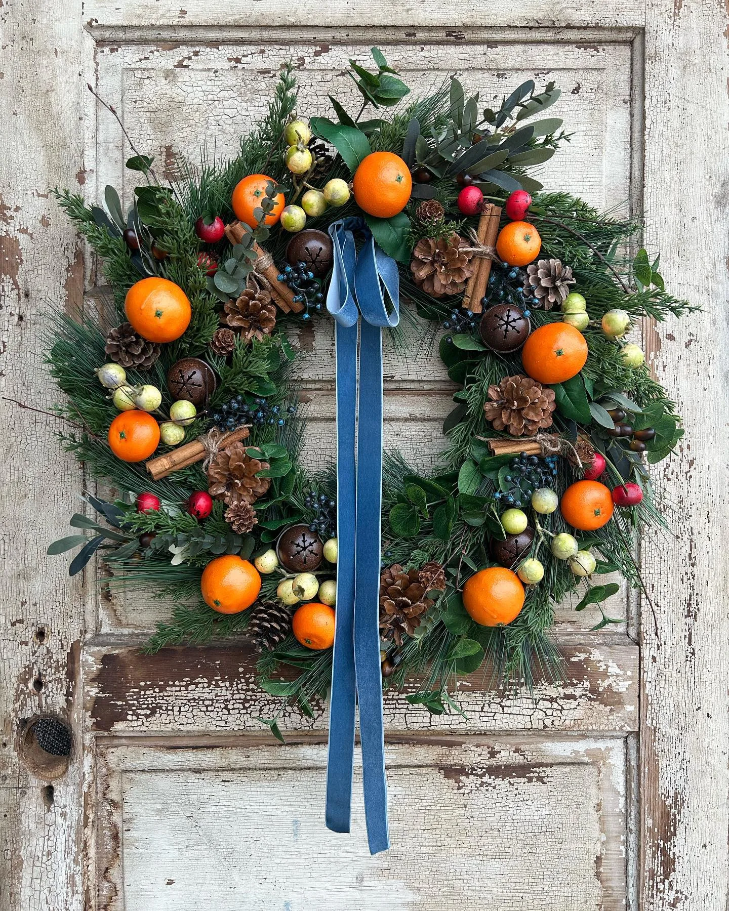 A festive wreath decorated with oranges, pinecones, berries, and green foliage, adorned with a blue ribbon, hangs on a rustic wooden door. Discover how to make your own fall wreaths with natural materials and scents to bring this charming touch to your home.