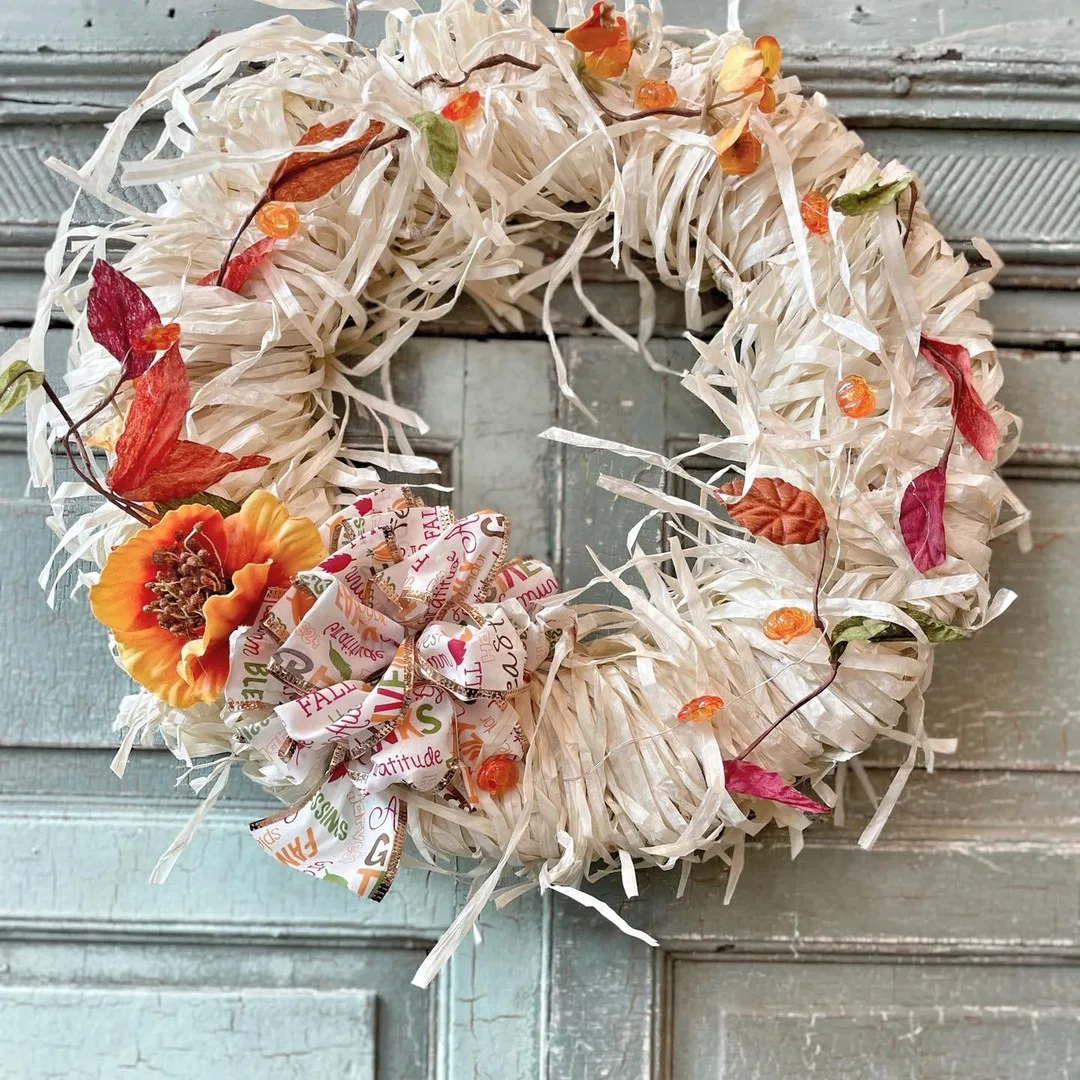 A wreath made of white shredded material, adorned with autumn leaves, a large orange flower, small orange decorations, and a ribbon with words like "fall" and "gratitude," hangs on a rustic wooden door. Learn *how to make your own fall wreaths with natural materials and scents* for an extra touch of seasonal charm.