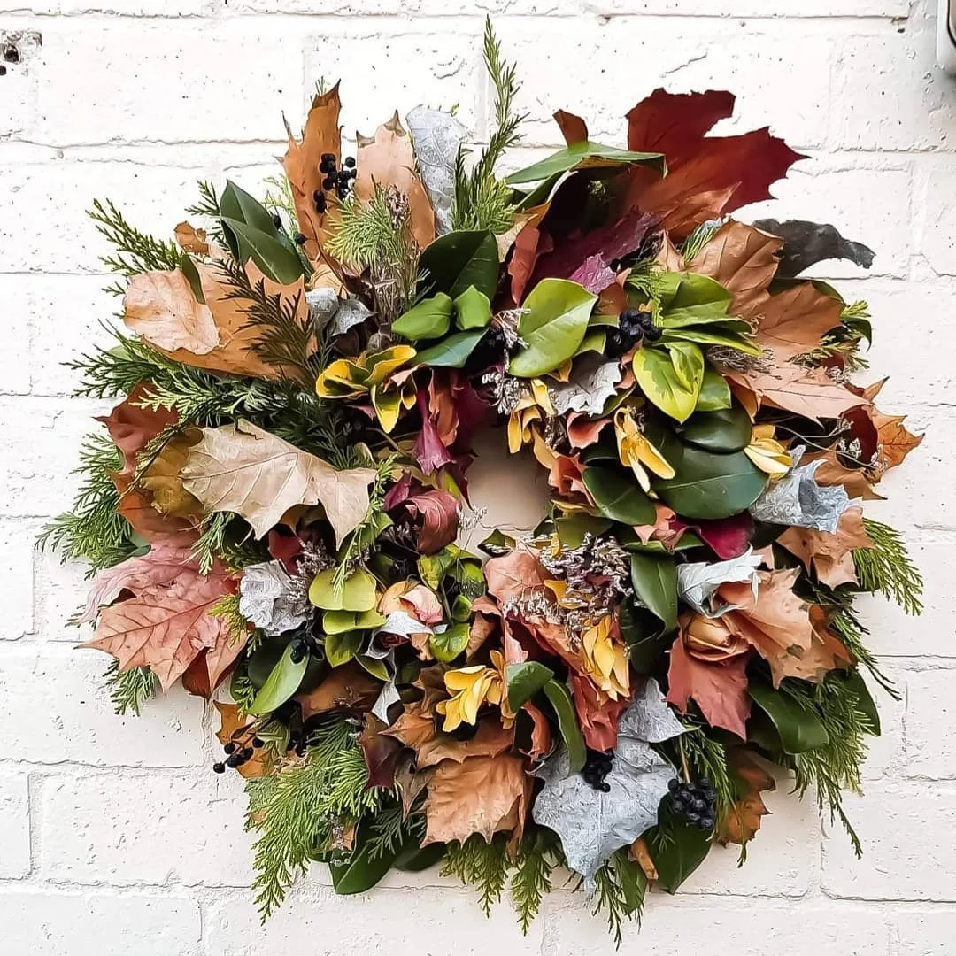 A decorative wreath made of autumn leaves and greenery is hung on a white brick wall, showcasing how to make your own fall wreaths with natural materials and scents.