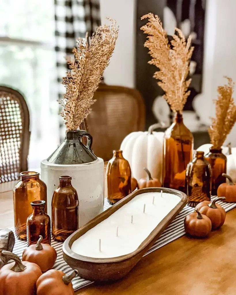A fall-themed table centerpiece featuring amber glass bottles, dried grasses, mini pumpkins, white gourds, and a wooden tray with a large candle exuding rustic romance. Captivating fall centerpieces with the warmth of candlelight.