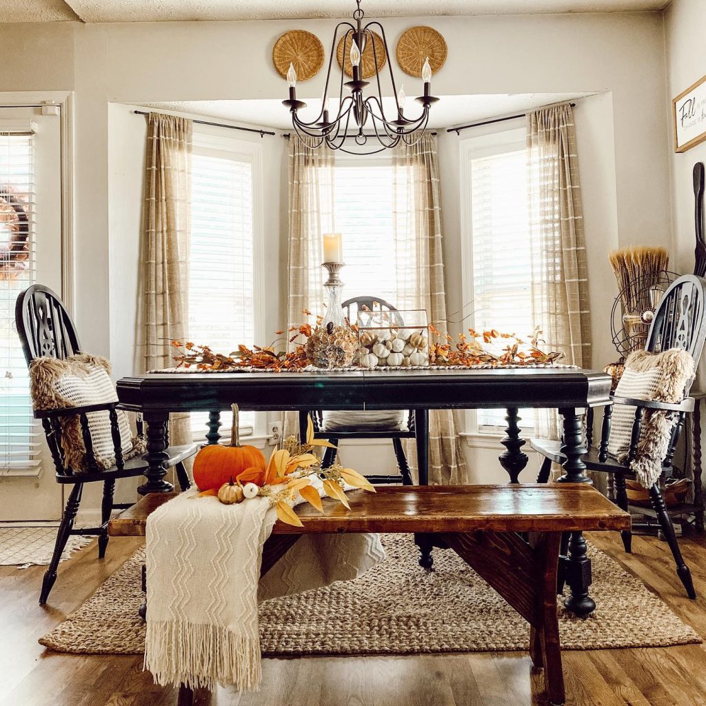 A dining room features a black table adorned with autumn decor, surrounded by black chairs. A bench with a pumpkin and blanket sits in front, offering one of the many 4 Fall Creative Table Decor Ideas Using Branches and Leaves. Beige curtains and a chandelier grace the background.