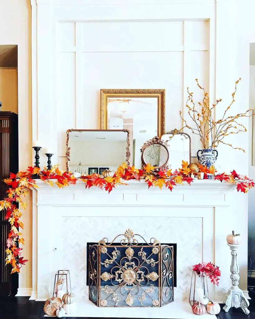 A white fireplace mantle is adorned with rich autumnal hues, featuring a fall leaves garland, pumpkins, candles, a mirror, and a vase with branches on top. The ornate fireplace screen enhances the cozy fall decor ambiance.