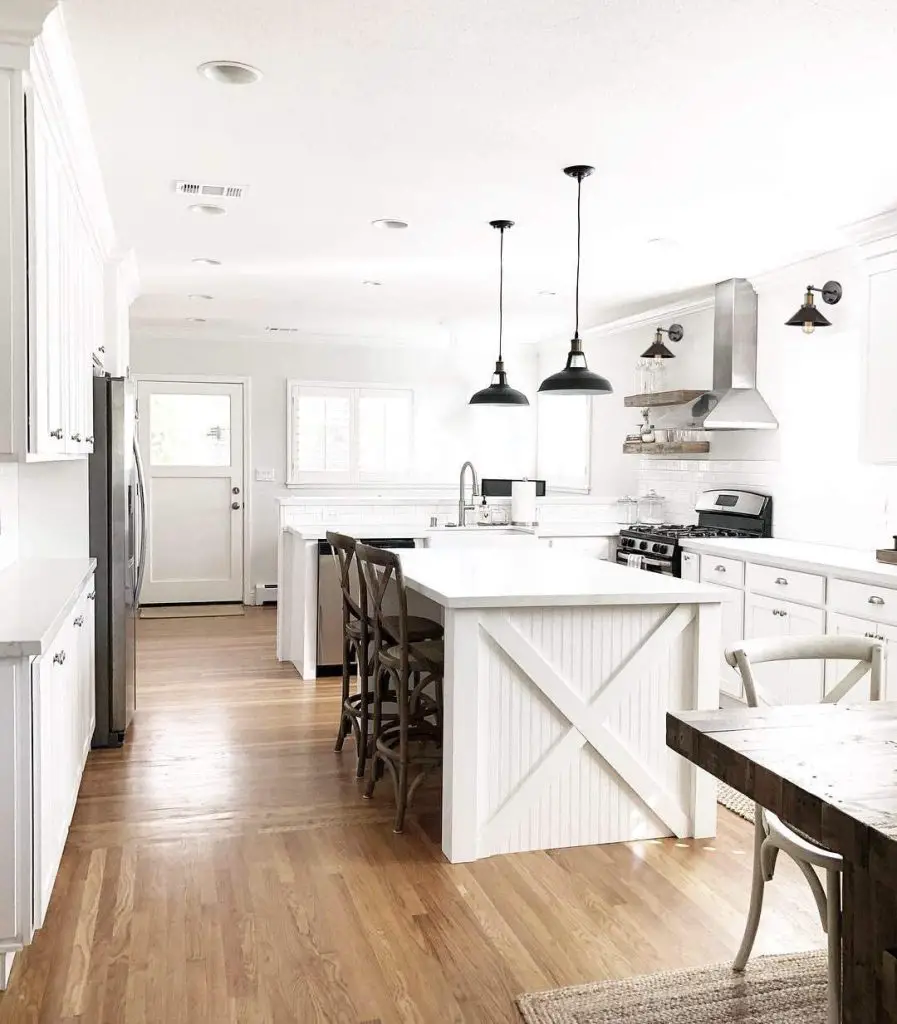 Bright, modern kitchen with white cabinets, stainless steel appliances, an island with barstools, and wood flooring. Enchant your home with rustic charm by adding the perfect industrial barn light to complement the pendant lighting.
