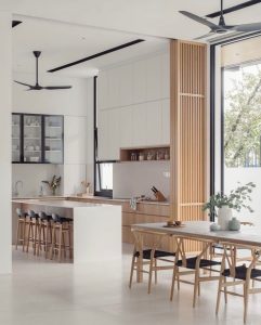 Modern kitchen and dining area with white and wood accents, black bar stools, overhead ceiling fans, and large windows. The kitchen includes open shelving, a countertop with stools, and a dining table. Japandi-inspired design ideas create a functional and beautiful space.