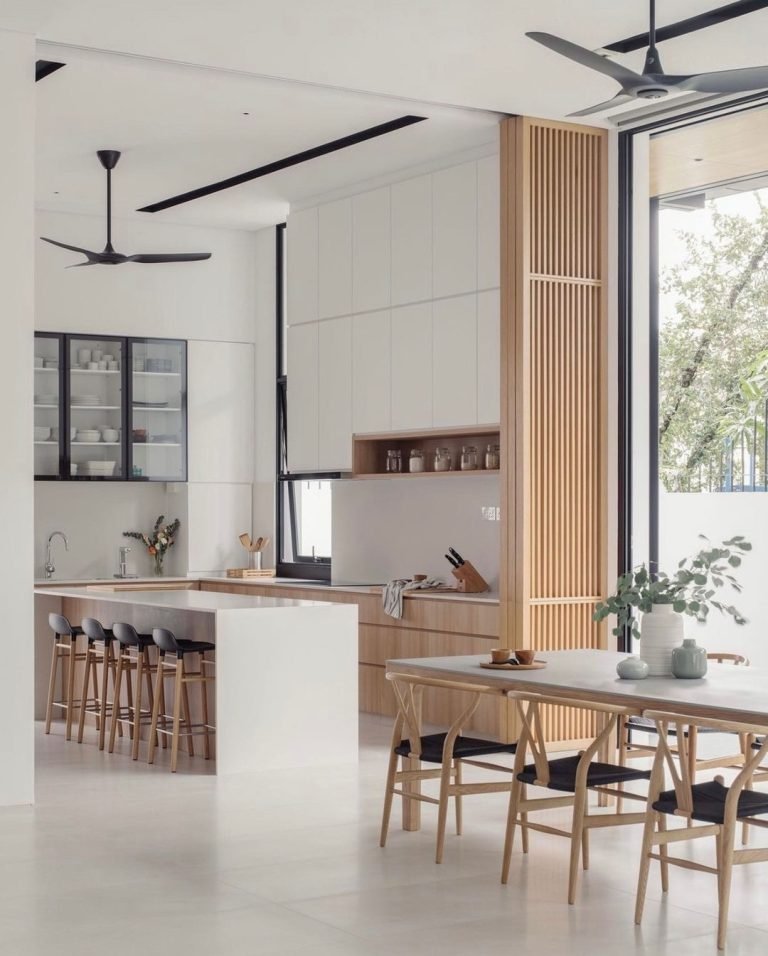 Modern kitchen and dining area with white and wood accents, black bar stools, overhead ceiling fans, and large windows. The kitchen includes open shelving, a countertop with stools, and a dining table. Japandi-inspired design ideas create a functional and beautiful space.