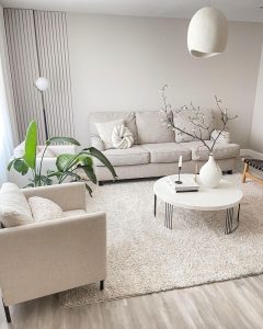 A minimalistic living room embodies mindfulness in Japandi decor, featuring a beige sofa, armchair, white round coffee table with a vase, and green plants on a light-colored rug against pale walls.
