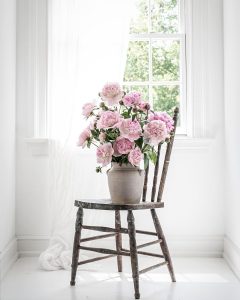 A rustic wooden chair with a white vase of pink peonies is placed in front of a window with sheer curtains, allowing natural light to illuminate the scene, showcasing one of the 15 ways to create a stunning romantic bedroom with pink peonies.