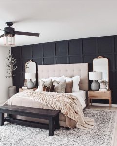 A bedroom with a beige tufted headboard, black accent wall, and wooden furniture demonstrates how to style with the perfect furniture pieces for a transitional bedroom. It includes a bed, two side tables with lamps, a bench at the foot of the bed, and decorative plants on both sides.