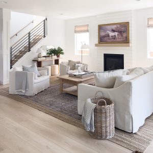 A transitional style living room with white walls, light wood flooring, a fireplace featuring a horse painting above, a beige sofa and armchairs, a wooden coffee table, a wicker basket with a blanket, and a staircase with a metal railing.