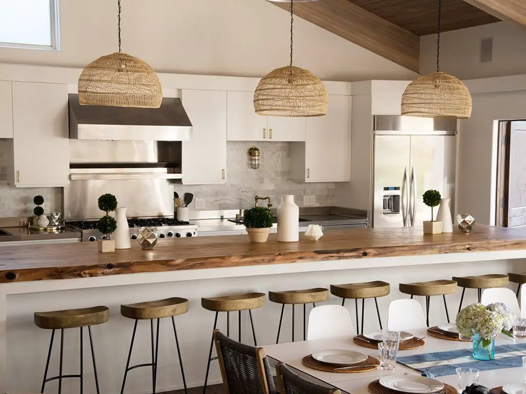 Modern kitchen with a long wooden island countertop, six stools, stainless steel appliances, and tropical wicker pendant lights. The island features decorative plants, canned jars, and a white vase, creating a serene oasis in your home.