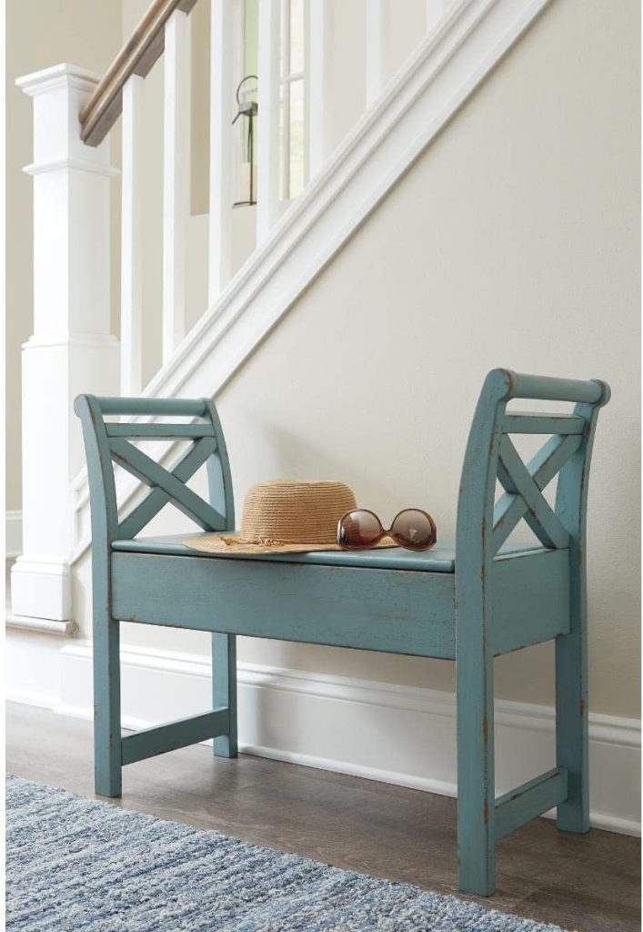 Entryway Elegance: A small blue wooden bench with a decorative backrest is placed beside a staircase, embodying rustic charm. A straw hat and a pair of sunglasses rest on the bench's seat, adding to its stylish appeal. The walls and staircase are painted white.