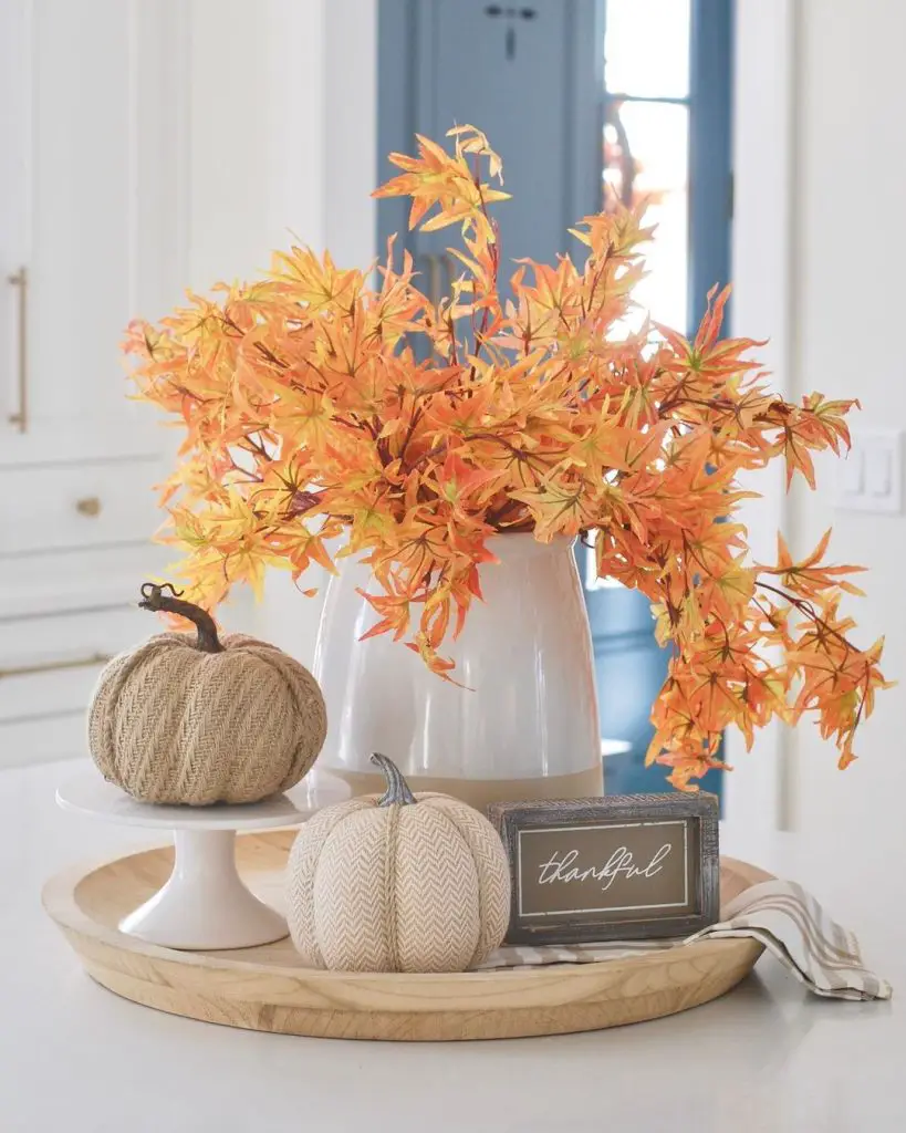 A white vase with orange fall foliage, two decorative pumpkins, and a framed "thankful" sign are displayed on a round wooden tray on a white countertop, embracing Hygge: Cozy Fall Decor Ideas for a Blissful Home.