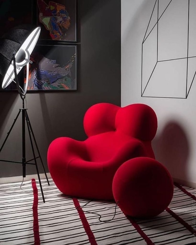 Red modern armchair with rounded shapes placed on a striped rug, next to a large floor lamp with a reflector and geometric wall art, illustrating perfect living room decor.