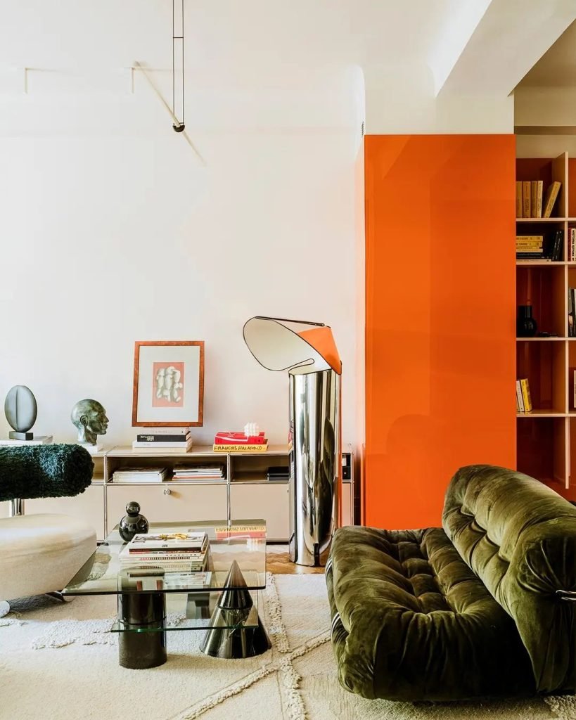 Modern living room with green velvet sofa, glass coffee table, and sculptural decor. Features a bold orange accent wall with built-in shelving and various decorative items and books. For enhanced living room decor, learn how to choose the best accent chair to perfectly complement the stylish ensemble.