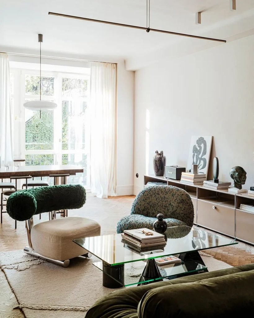 A modern living room with a glass coffee table, green and beige chairs, a dining table by the window, light-colored curtains, and minimalist decor including books and art pieces. Learn more in Living Room Decor: How to Choose the Best Accent Chair for your space.