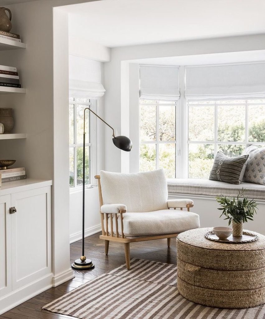 A cozy living room corner features a cushioned armchair, a round woven coffee table, a standing lamp, a striped rug, and tips on living room decor: how to choose the best accent chair. A built-in bookcase and window seat with pillows complete this well-lit space.