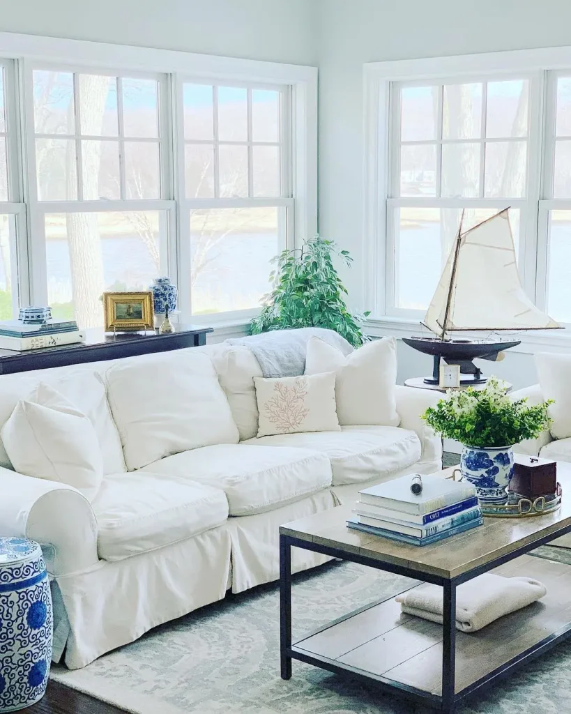 Bright living room with a white slipcover sofa showcasing how slipcover sofas enhance your living room's charm, nautical decor, coffee table with books and flowers, large windows, and a model sailboat on a side table.