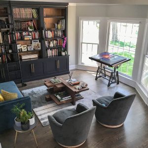 A spacious, sunlit living room embraces eclectic charm with a bookshelf, sofa, two armchairs, and a beautifully styled coffee table. A drafting table by the windows completes this inviting space.